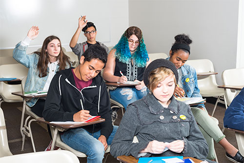 A classroom of six students at desks write in their notebooks. Two students in the back raise their hands. 